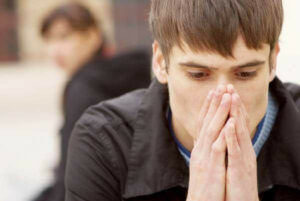 Image of young Caucasian man with hands over his nose and mouth contemplating whether or not to help a family member.